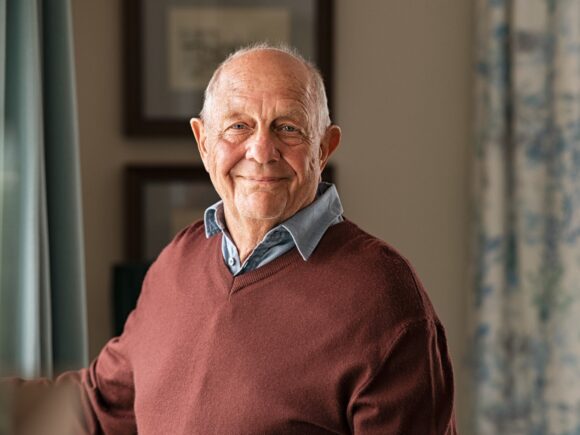 A balding older man stands by a living room window and smiles at the camera, he wears a blue shirt under a burgundy sweater.