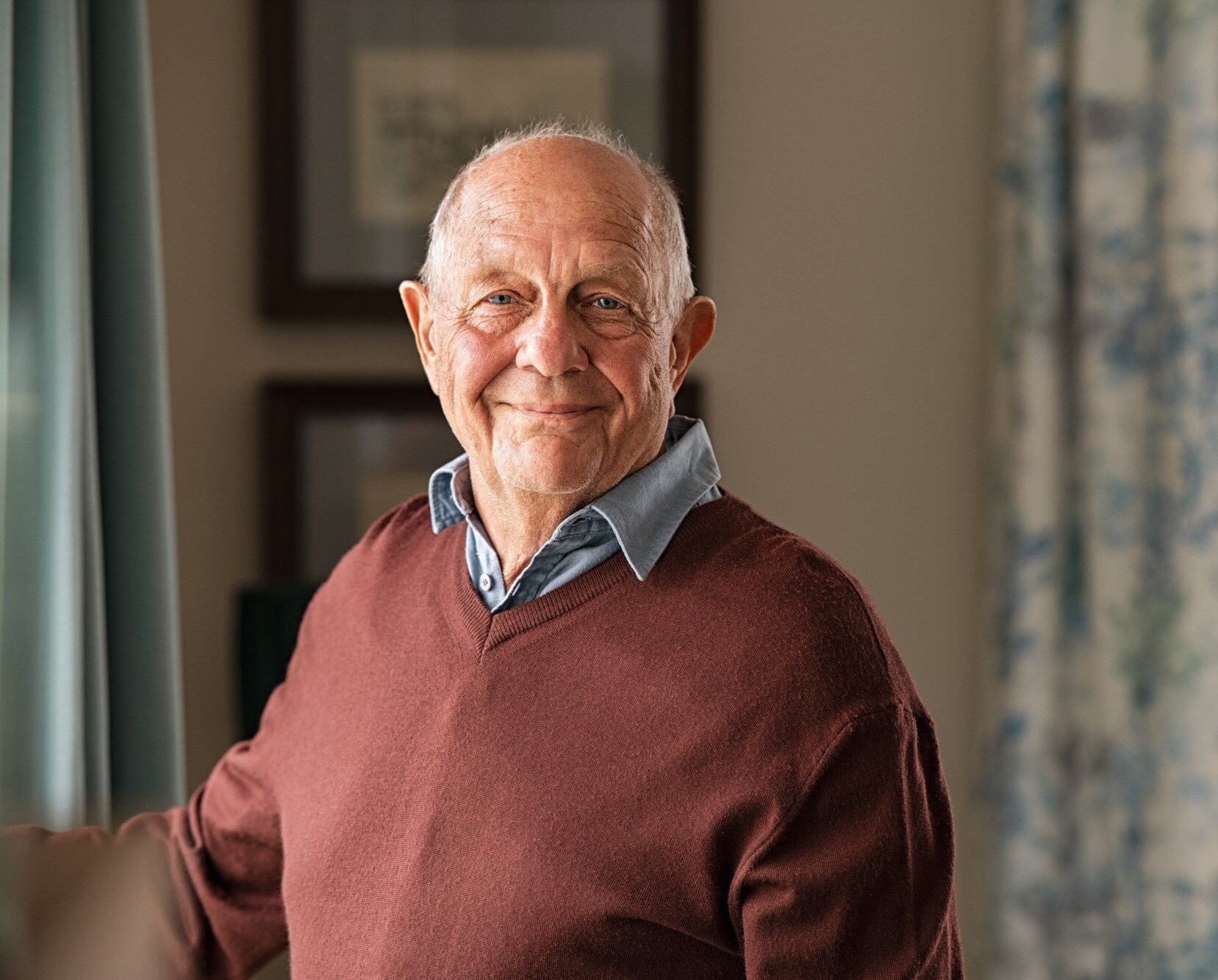 A balding older man stands by a living room window and smiles at the camera, he wears a blue shirt under a burgundy sweater.