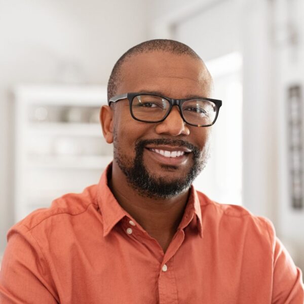 A man with shaved hair and a short black beard smiles at the camera, he wears thick rimmed glasses and an orange button-down shirt.