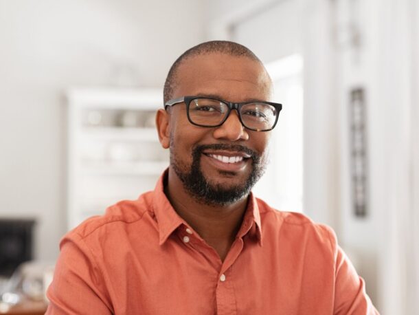 A man with shaved hair and a short black beard smiles at the camera, he wears thick rimmed glasses and an orange button-down shirt.