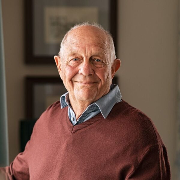 A balding older man stands by a living room window and smiles at the camera, he wears a blue shirt under a burgundy sweater.