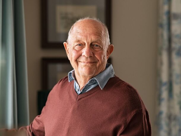 A balding older man stands by a living room window and smiles at the camera, he wears a blue shirt under a burgundy sweater.