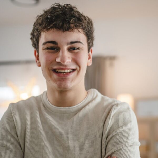 A teenage man with short brown curly hair smiles broadly at the camera, he wears a beige long sleeved top.