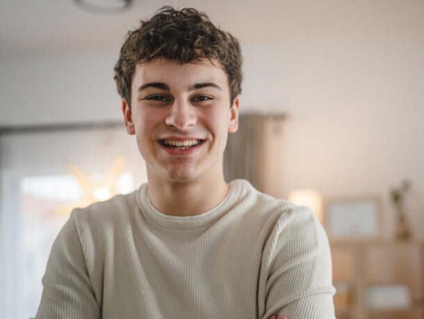 A teenage man with short brown curly hair smiles broadly at the camera, he wears a beige long sleeved top.