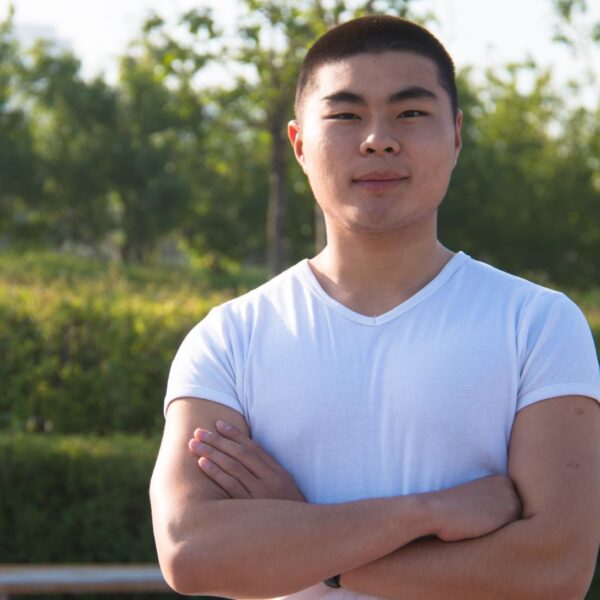 A young man with short black hair stands outside wearing a white v-neck t-shirt, his arms are folded and he wears a mild smile.