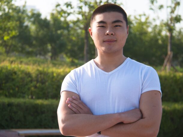 A young man with short black hair stands outside wearing a white v-neck t-shirt, his arms are folded and he wears a mild smile.