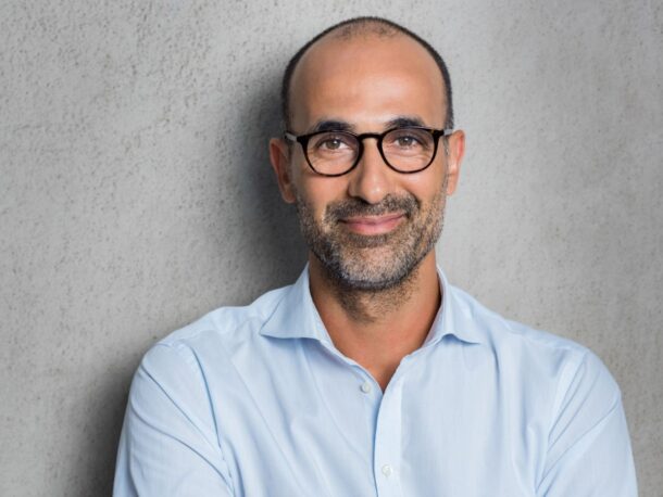 A man with very short greying, black hair with a very short beard smiles at the camera, he's wearing black rimmed glasses and a pale blue button-down shirt.