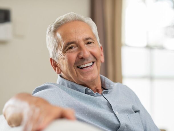 An older man sits on a sofa smiling, his arm is stretched along the back of the sofa towards the camera.