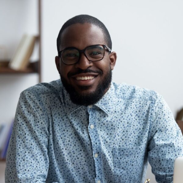 A young man with very short black hair and a short black beard smiles at the camera, he's wearing glasses with thick frames and a floral patterned, blue button-down shirt.