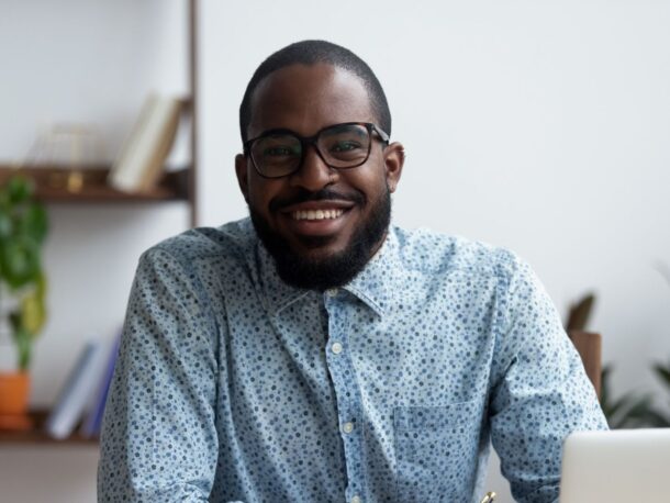 A young man with very short black hair and a short black beard smiles at the camera, he's wearing glasses with thick frames and a floral patterned, blue button-down shirt.