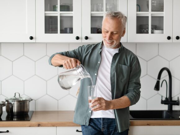 A man stands in a kitchen leaning against a worktop while pouring water from a jug into a glass.