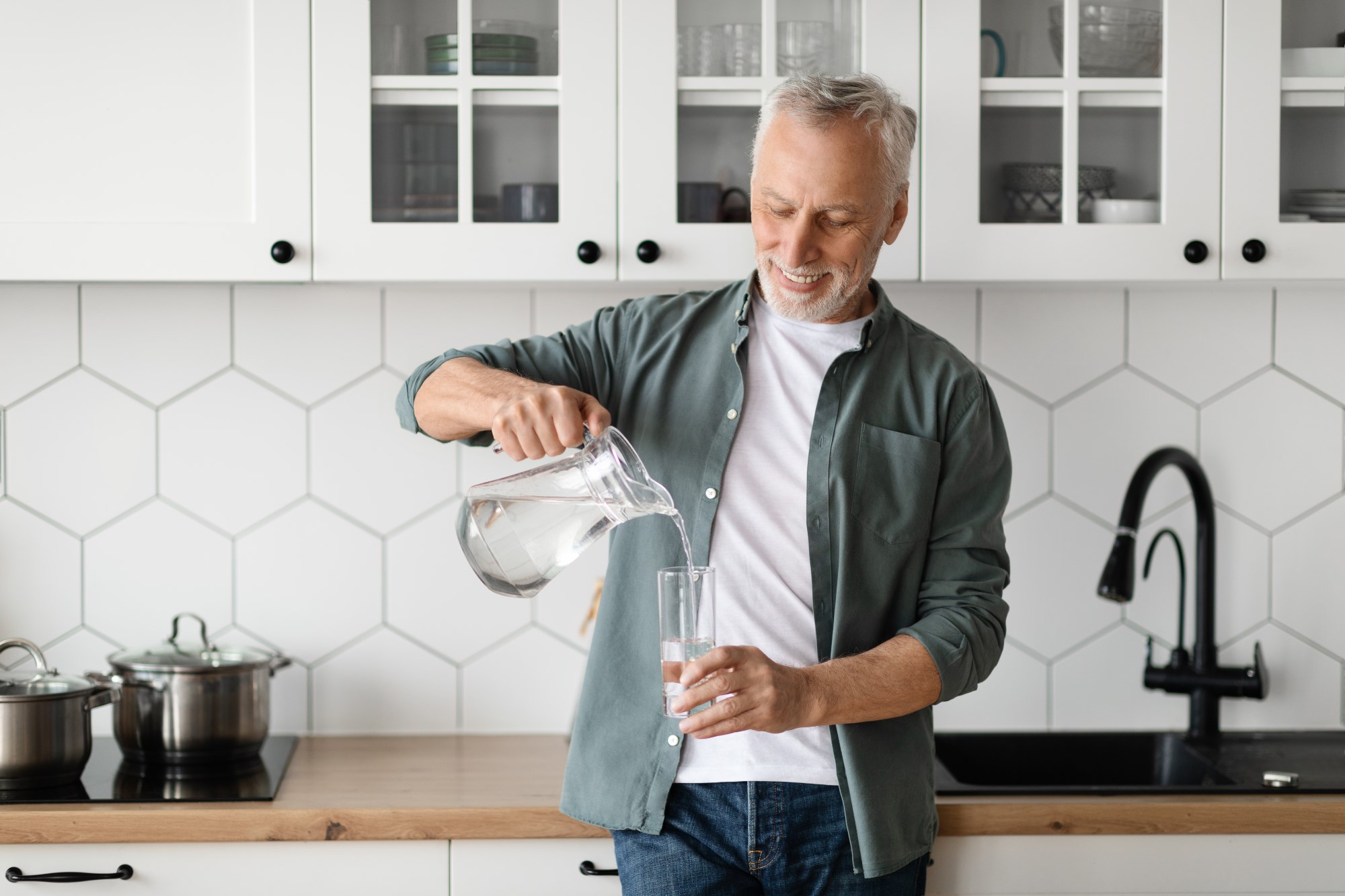 A man stands in a kitchen leaning against a worktop while pouring water from a jug into a glass.