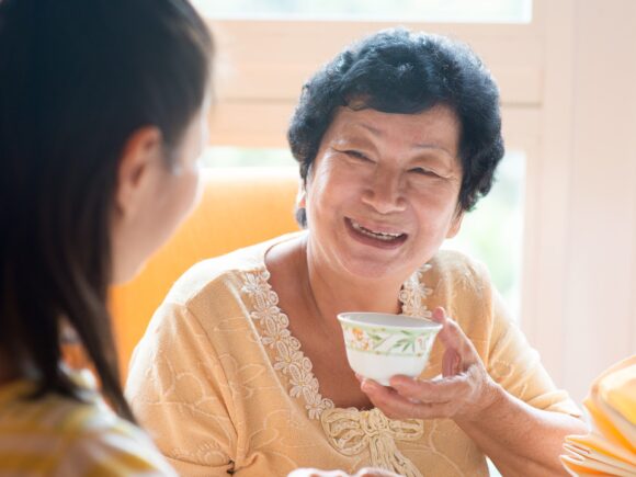 An older Asian woman holds up a teacup as she laughs with a younger woman in the foreground.