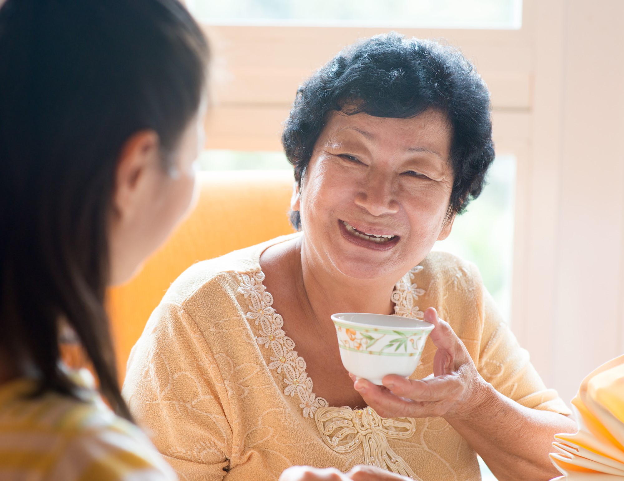 An older Asian woman holds up a teacup as she laughs with a younger woman in the foreground.