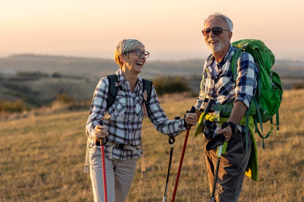 An older couple standing on a hill at sunset, they are holding hiking poles and have a large rucksack. They are both smiling and laughing.