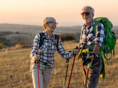 An older couple standing on a hill at sunset, they are holding hiking poles and have a large rucksack. They are both smiling and laughing.