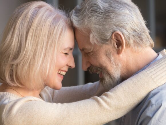 An older couple embrace, smiling they touch foreheads as they wrap their arms around each other.