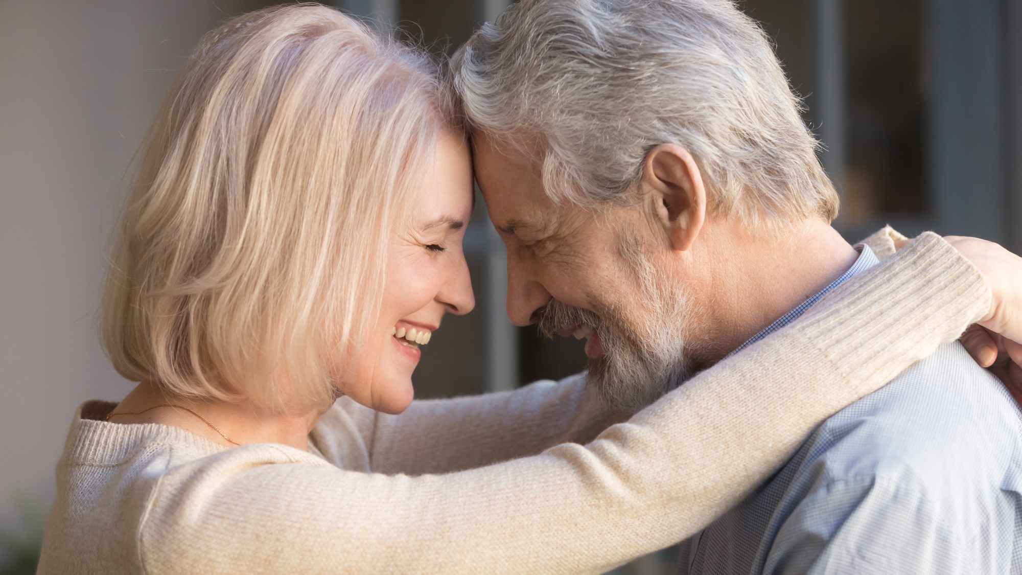 An older couple embrace, smiling they touch foreheads as they wrap their arms around each other.
