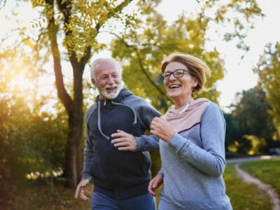 An older couple run through a park side by side, smiling and laughing.
