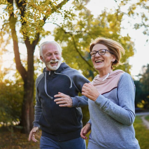 An older couple run through a park side by side, smiling and laughing.