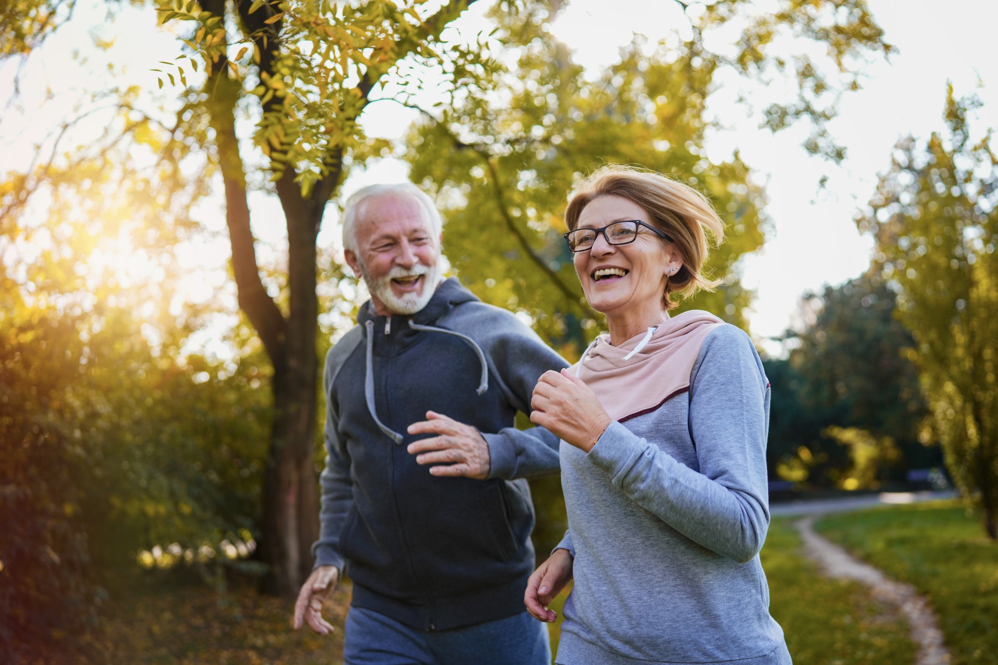 An older couple run through a park side by side, smiling and laughing.