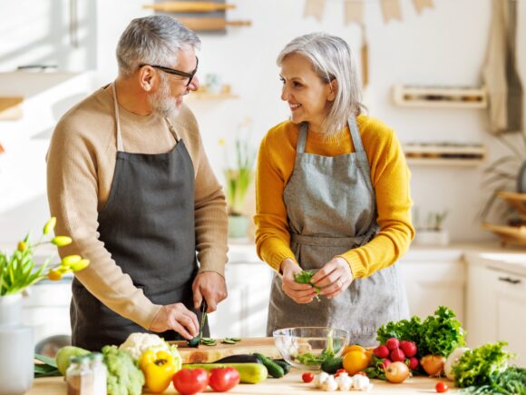 An older couple smile as they talk to each other and prepare vegetables on a kitchen island.