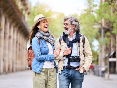 A middle aged couple walk arm-in-arm down an avenue laughing, they appear to be on holiday.