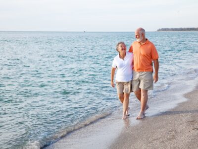 An older couple walk along the shoreline with the waves lapping at their bare feet. They both wear long beige shorts and polo shirts and are laughing as if sharing a joke.