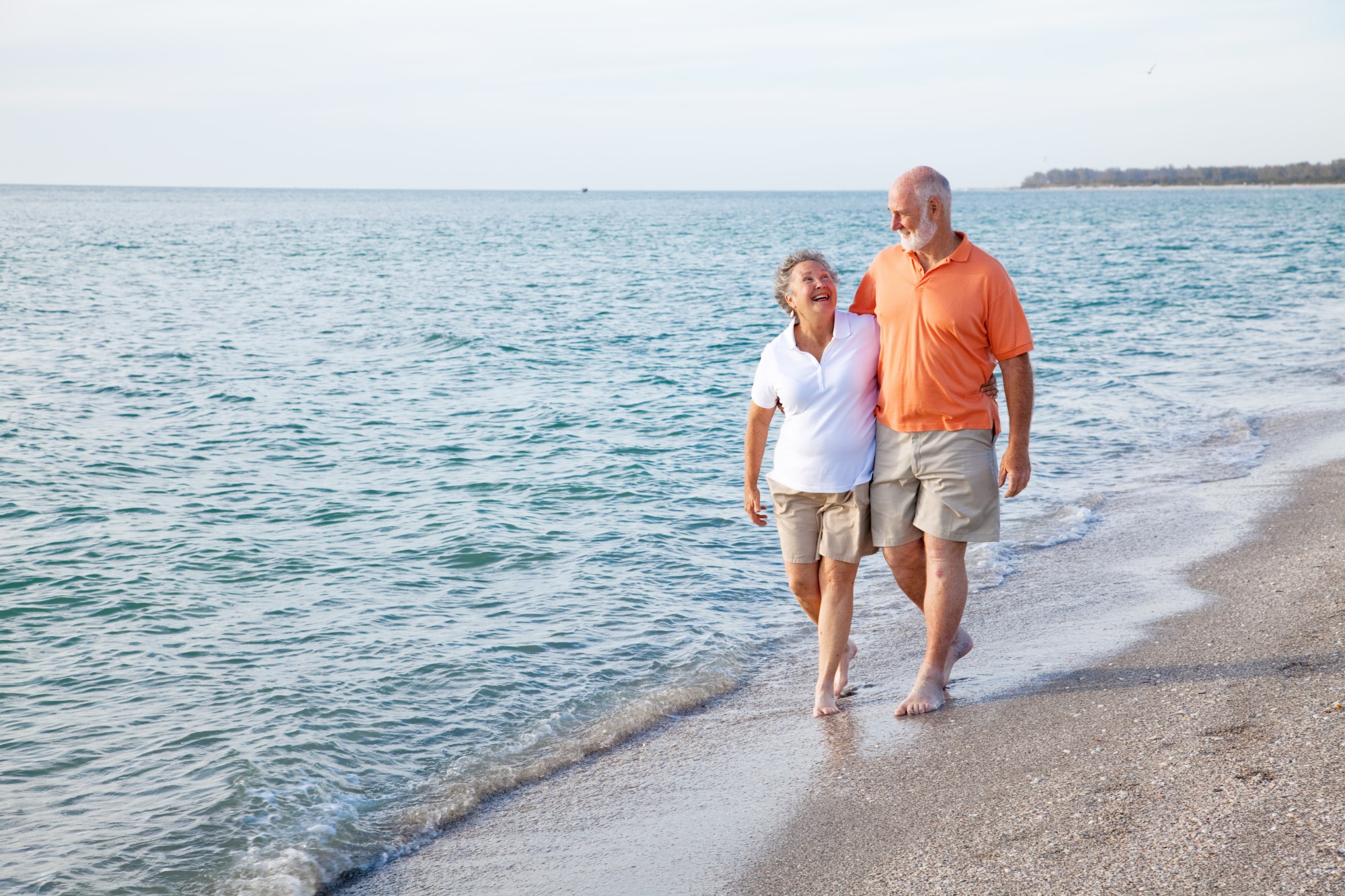 An older couple walk along the shoreline with the waves lapping at their bare feet. They both wear long beige shorts and polo shirts and are laughing as if sharing a joke.