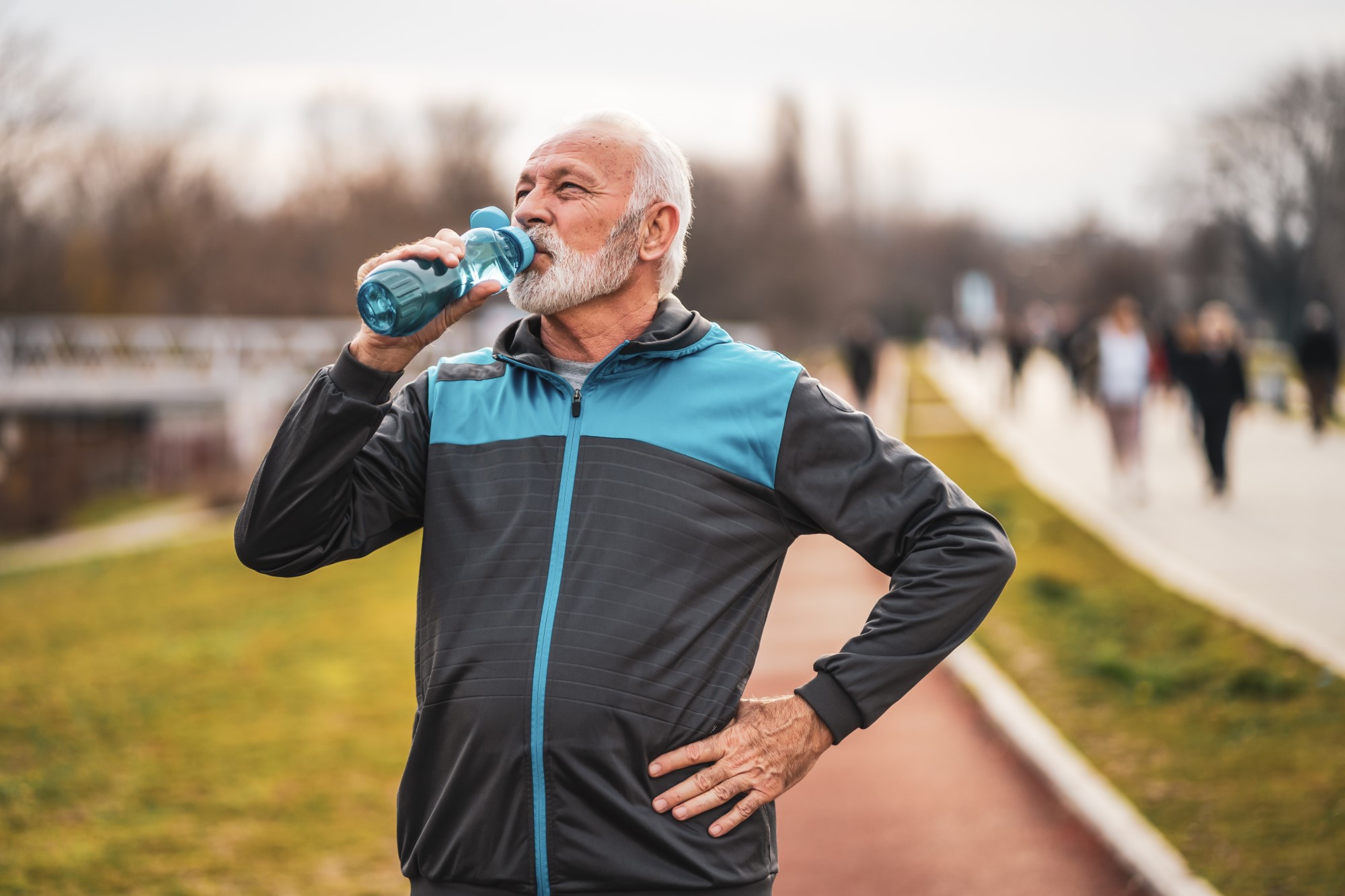An older man drinks from a water bottle, he appears to have just finished running in the park.