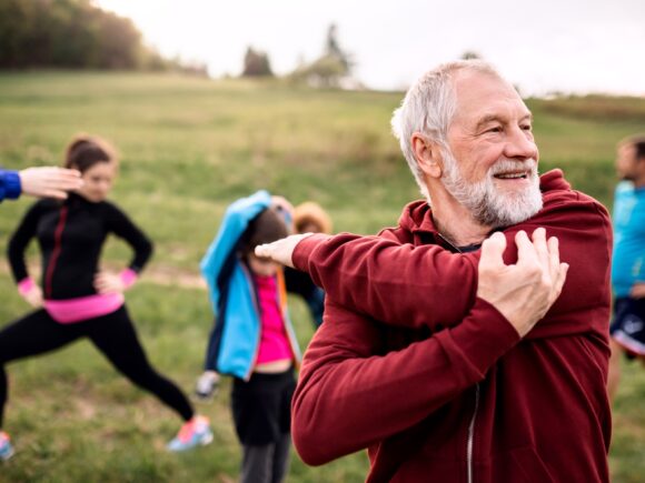 An older man doing some stretching exercises outside, behind him a group of people of various ages also stretch.