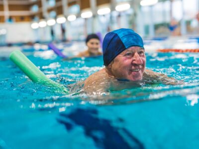 An older man swims length in an indoor swimming pool with the aid of a foam pool noodle, he is wearing a swimming cap and smiling.