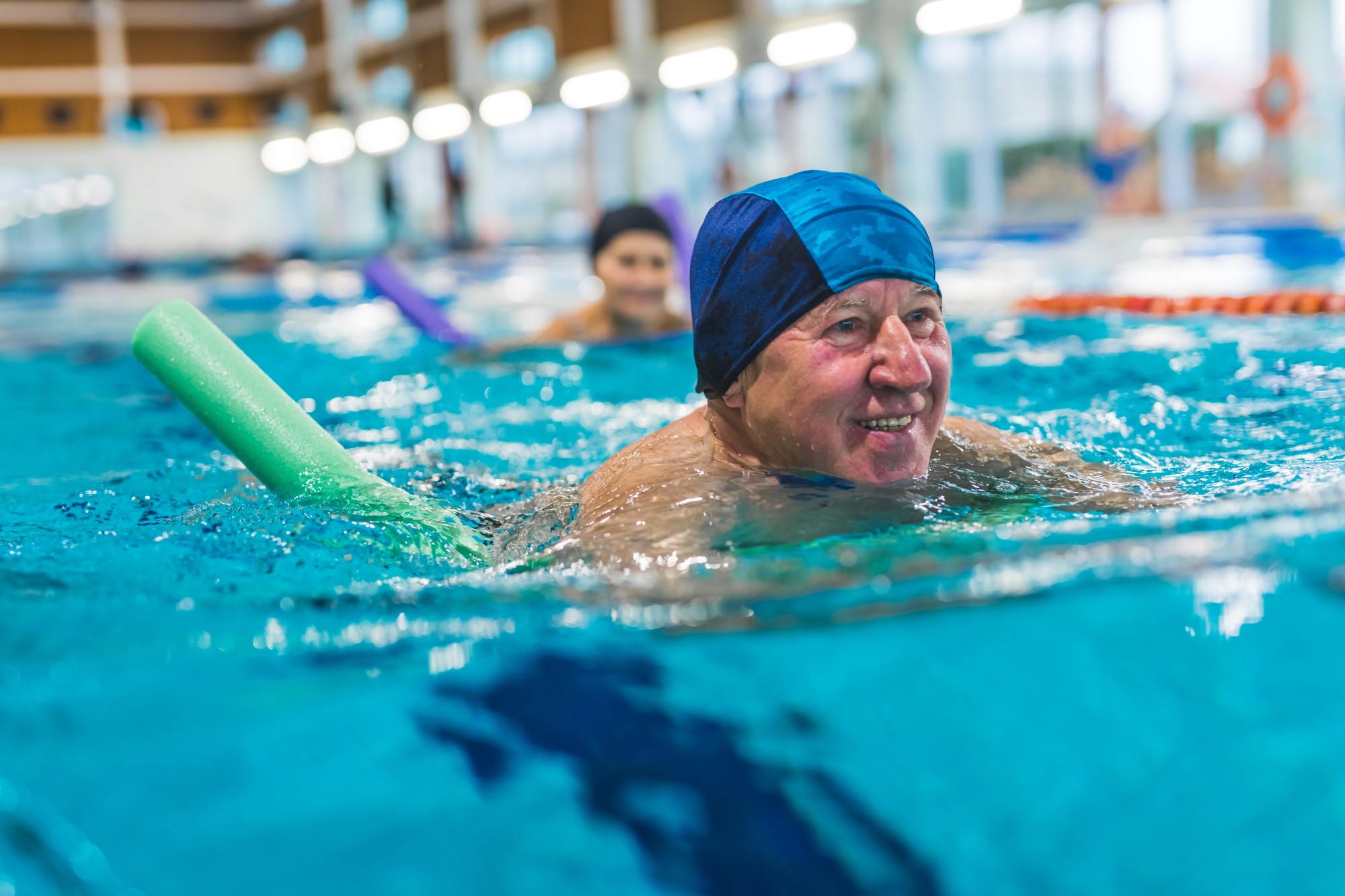 An older man swims length in an indoor swimming pool with the aid of a foam pool noodle, he is wearing a swimming cap and smiling.