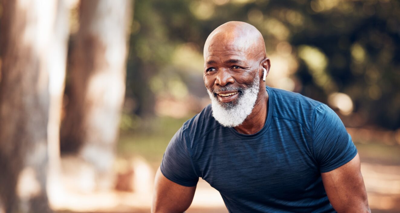 An older man with a white beard takes a break from running, he wears earbuds and leans forward with a hand on top of his thigh.