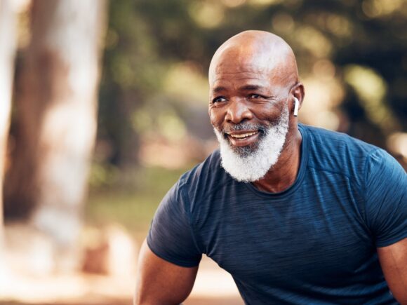 An older man with a white beard takes a break from running, he wears earbuds and leans forward with a hand on top of his thigh.
