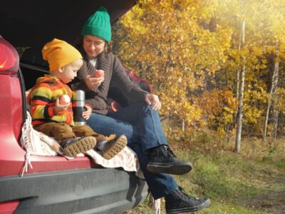 A parent and their young child sit in the boot of their car with a flask as they eat apples, they wear woolly hats and the leaves are turning autumn gold behind them.