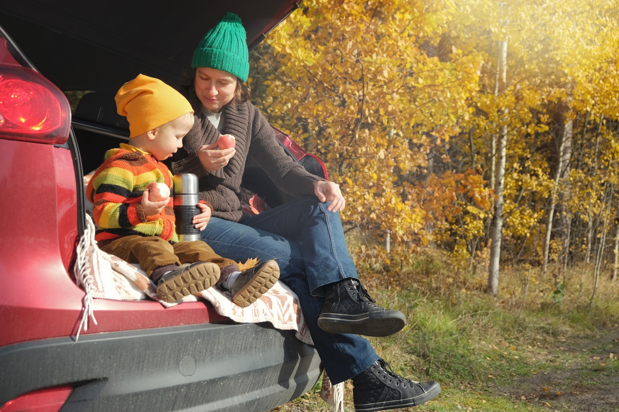 A parent and their young child sit in the boot of their car with a flask as they eat apples, they wear woolly hats and the leaves are turning autumn gold behind them.