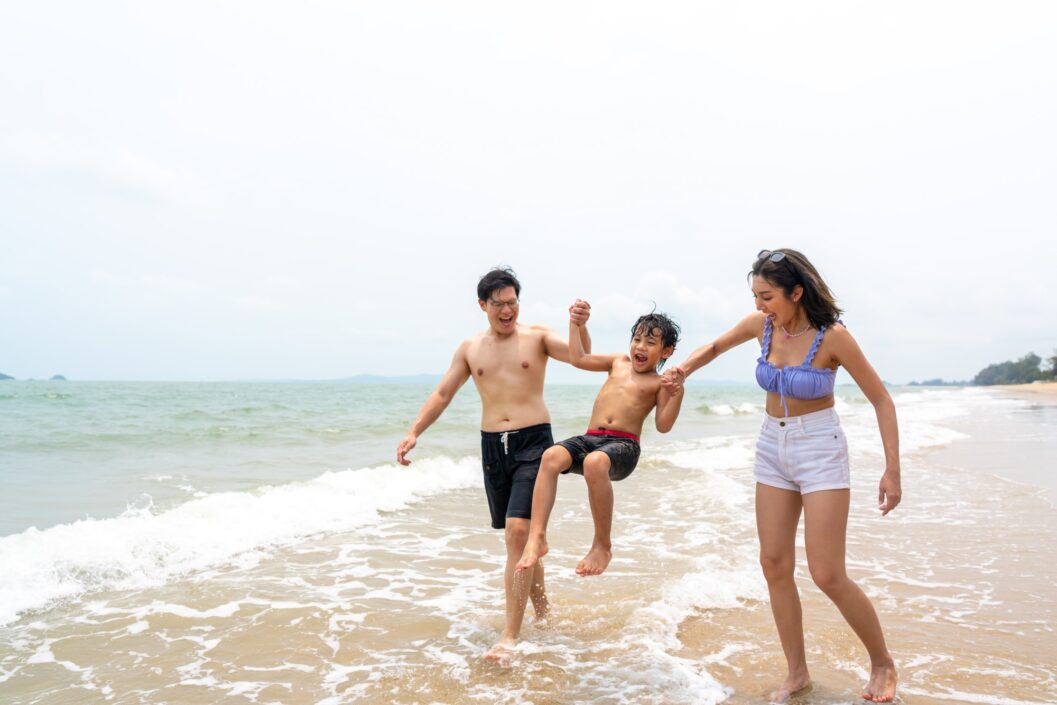 A couple swing their child between them as they walk through the surf on the beach. The parents and boy are all laughing.