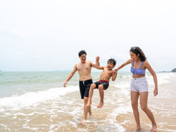 A couple swing their child between them as they walk through the surf on the beach. The parents and boy are all laughing.