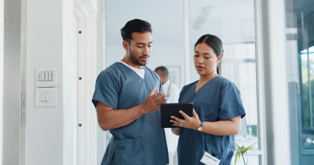 Two healthcare professionals stand in a hospital ward discussing notes on a tablet one of them is holding.