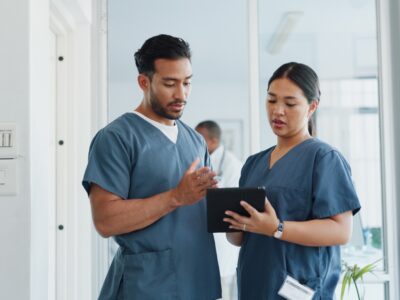 Two healthcare professionals stand in a hospital ward discussing notes on a tablet one of them is holding.