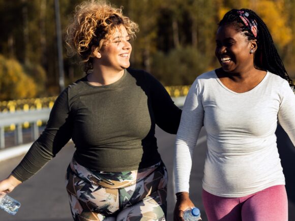 Two women wearing exercise clothes and holding water bottles walk down a road smiling and laughing, they appear to have just finished exercising.