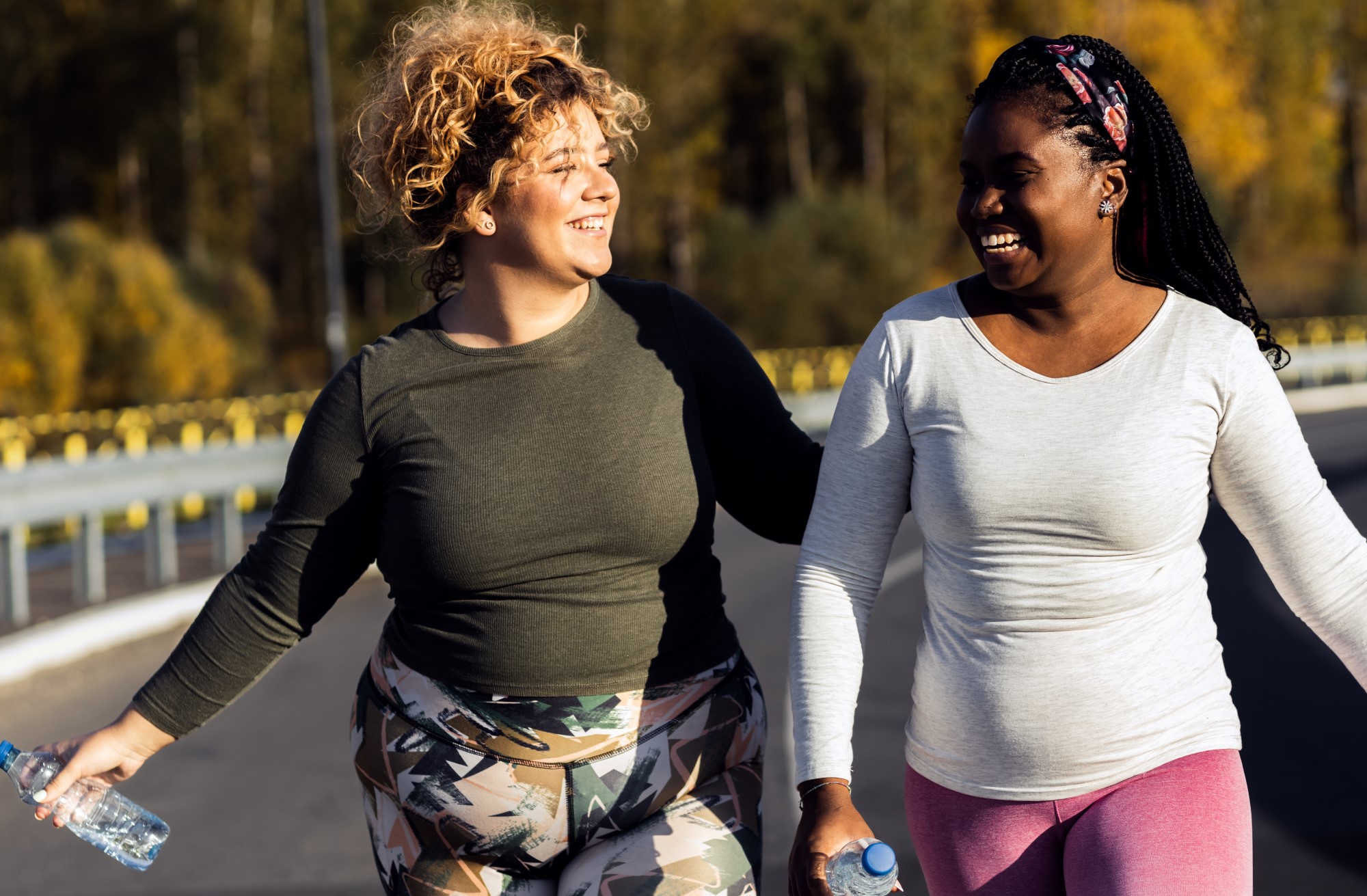 Two women wearing exercise clothes and holding water bottles walk down a road smiling and laughing, they appear to have just finished exercising.