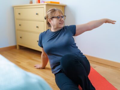 A middle aged lady, her brown hair in a bun, wearing glasses, a t-shirt and leggings performs a Pilates pose. She is seated with her legs outstretched, twisting to her left on a yoga mat in a bedroom.