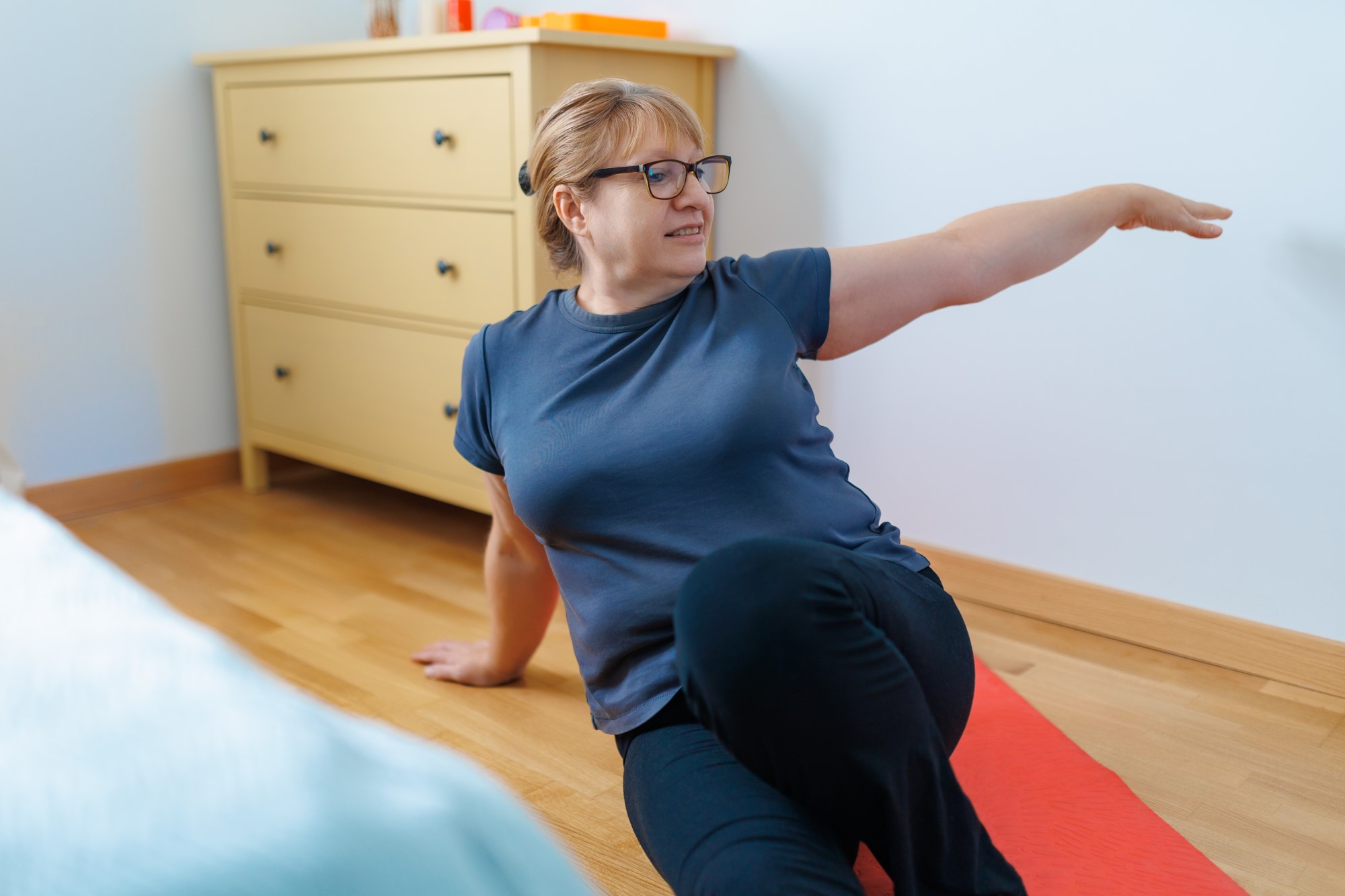 A middle aged lady, her brown hair in a bun, wearing glasses, a t-shirt and leggings performs a Pilates pose. She is seated with her legs outstretched, twisting to her left on a yoga mat in a bedroom.
