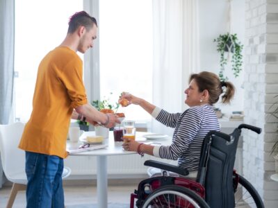 A woman in a wheelchair sits at a dining table taking a croissant from a plate offered by a young man in casual clothes, they could be mother and son.