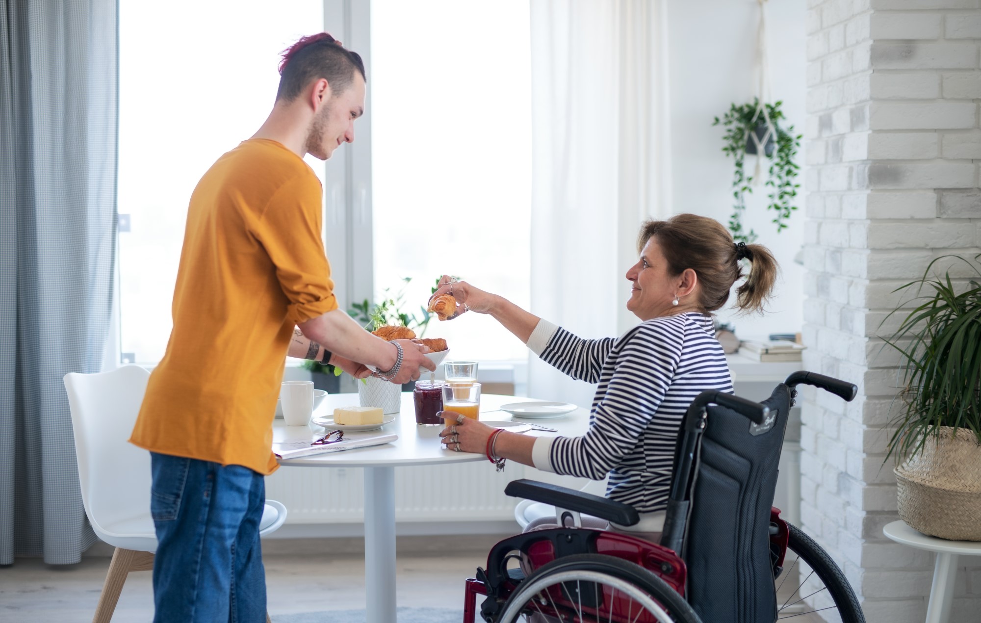 A woman in a wheelchair sits at a dining table taking a croissant from a plate offered by a young man in casual clothes, they could be mother and son.