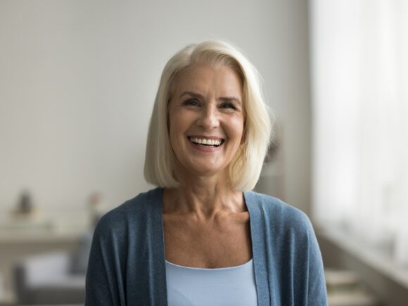 An older woman with a long light blonde bob and brown hair smiles wildly, she is wearing a blue cardigan.