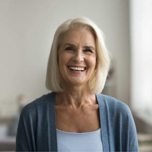 An older woman with a long light blonde bob and brown hair smiles wildly, she is wearing a blue cardigan.