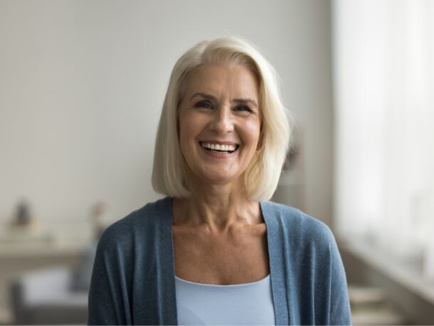 An older woman with a long light blonde bob and brown hair smiles wildly, she is wearing a blue cardigan.